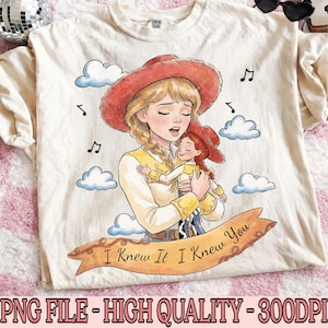 May include: A cream-colored t-shirt featuring a graphic illustration of a blonde character in a red cowboy hat holding a smaller doll version of themselves. The design includes blue clouds, musical notes, and a banner with the text I Knew It I Knew You.