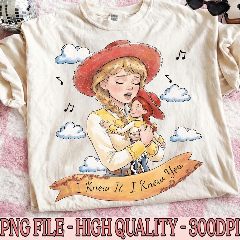 May include: A cream-colored t-shirt featuring a graphic illustration of a blonde character in a red cowboy hat holding a smaller doll version of themselves. The design includes blue clouds, musical notes, and a banner with the text I Knew It I Knew You.