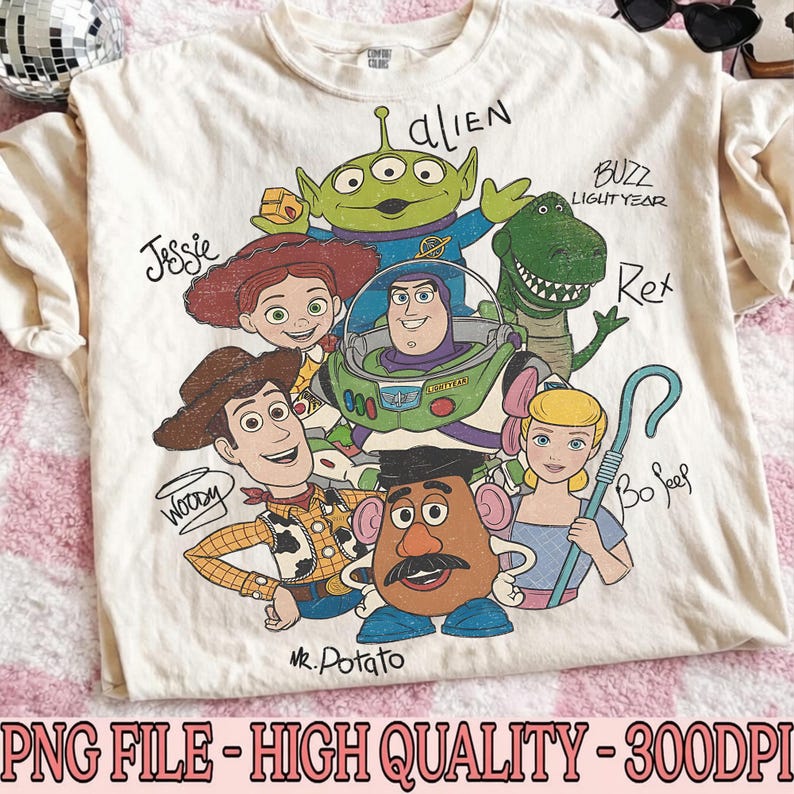 Puede incluir: Una camiseta de color crema con un estampado gr&aacute;fico de personajes de Toy Story, incluyendo a Woody, Buzz Lightyear, Jessie, Rex, Bo Peep, Mr. Potato Head y un alien&iacute;gena. Los nombres de cada personaje est&aacute;n escritos en una fuente manuscrita.