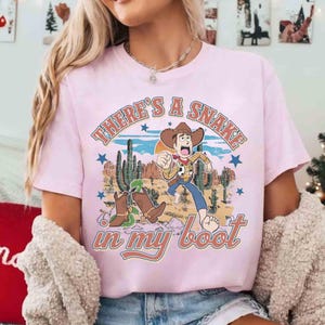 Peut inclure: T-shirt rose clair avec un motif de dessin anim&eacute; d'un cow-boy courant dans un paysage d&eacute;sertique. Le texte "THERE'S A SNAKE in my boot" est imprim&eacute; au-dessus et en dessous du motif. Le t-shirt a des manches courtes.