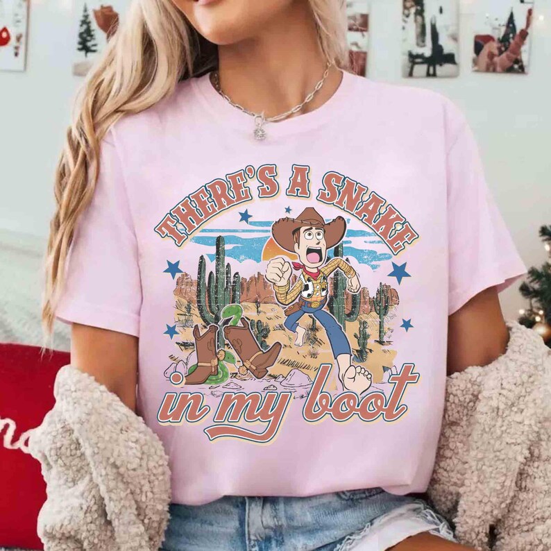Peut inclure: T-shirt rose clair avec un motif de dessin anim&eacute; d'un cow-boy courant dans un paysage d&eacute;sertique. Le texte "THERE'S A SNAKE in my boot" est imprim&eacute; au-dessus et en dessous du motif. Le t-shirt a des manches courtes.