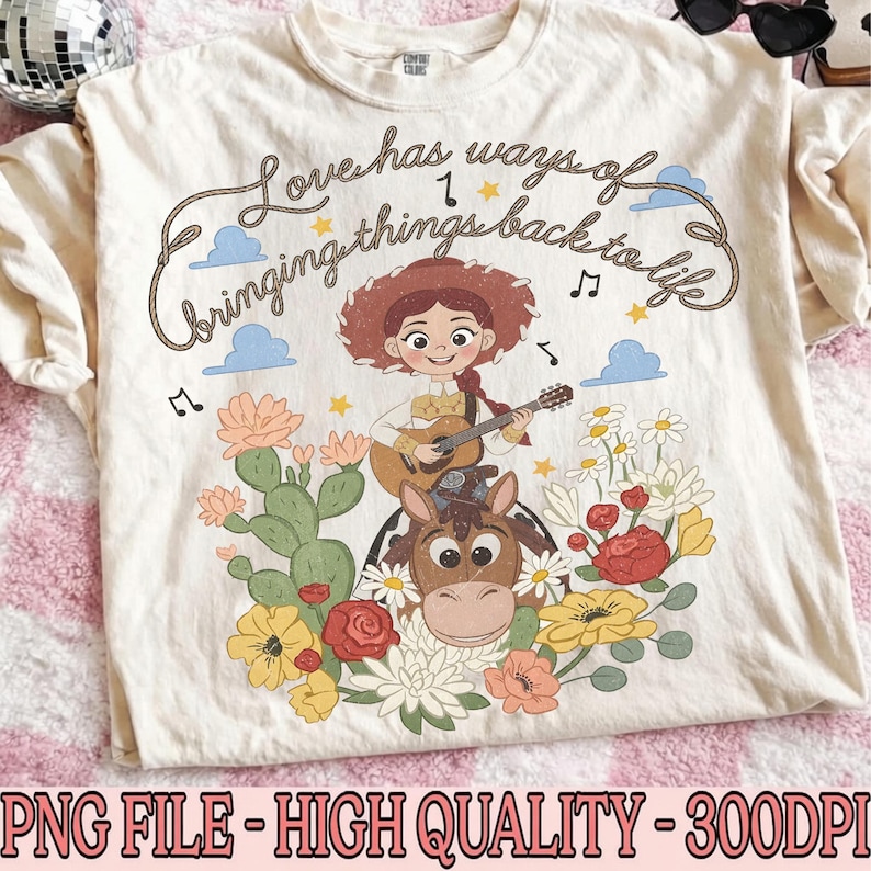 May include: A cream-colored t-shirt featuring a graphic of a cowgirl playing a guitar while riding a horse. The design includes the text "Love has ways of bringing things back to life" surrounded by cacti, colorful flowers, clouds, and musical notes.