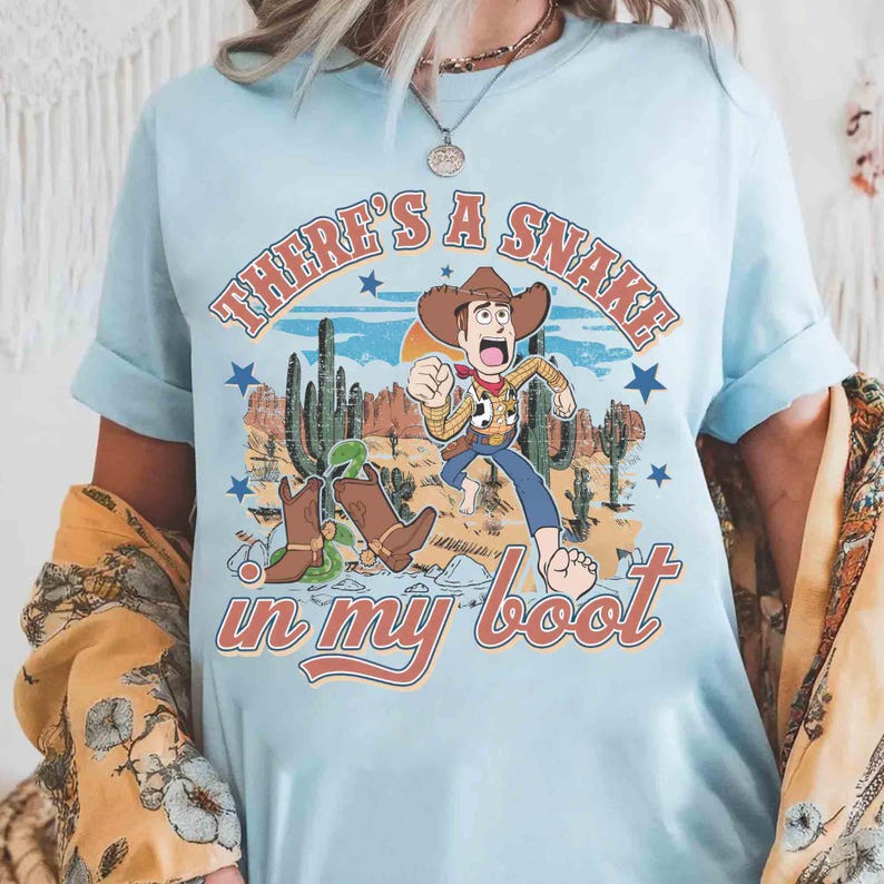 Peut inclure: T-shirt bleu clair avec une illustration de dessin anim&eacute; d'un cow-boy fuyant un serpent dans une botte. Le texte "THERE'S A SNAKE in my boot" est imprim&eacute; au-dessus et en dessous de l'illustration en orange et rouge.