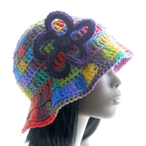 May include: A colorful crocheted bucket hat with a large purple flower on the side. The hat is made with a variety of colors, including red, orange, yellow, green, blue, and purple.