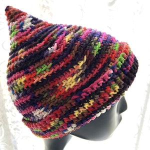 May include: A colorful, handmade crochet hat with a pointed top. The hat features a vibrant mix of colors including red, pink, yellow, green, and purple. The hat is displayed on a black mannequin head.