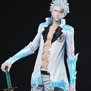 Peut inclure: Figurine détaillée d'un personnage aux cheveux blancs hérissés, vêtu d'un manteau blanc avec des accents bleus glacés. La figurine tient une épée et a un physique musclé. La figurine est de Tanuki Figures.