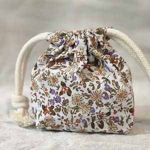 Puede incluir: Una pequeña bolsa con cordón con un estampado floral en blanco, marrón, naranja y morado. La bolsa tiene un cierre de cordón de cuerda blanca. La bolsa probablemente está hecha de tela y es adecuada para objetos pequeños.