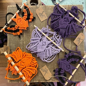 May include: Five macrame wall hangings in various colors and patterns. The hangings are made with cotton cord and feature geometric designs. The hangings are attached to wooden dowels.