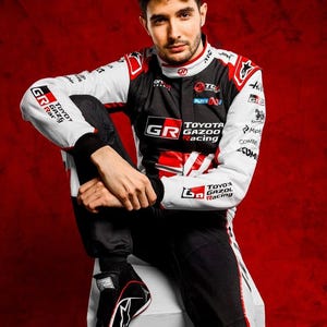 以下が含まれることがあります： トヨタ・ガズー・レーシングのロゴが入った、黒、白、赤のカラーリングのレーシングスーツ。長袖には様々なスポンサーロゴが施され、ハイカラーと黒のズボンが特徴です。デザインにはGRロゴが大きくあしらわれています。