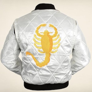 Puede incluir: Una chaqueta bomber de satén blanco acolchado con un gran escorpión amarillo bordado en la espalda. La chaqueta tiene un cuello y un dobladillo de canalé negro, mangas largas y un patrón de costuras en forma de diamante en toda la prenda.
