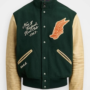 Pode incluir: Casaco universitário verde com mangas de couro bege. O casaco apresenta o texto "New England Run Club 1967" e um emblema alado. Possui gola, punhos e bainha canelados e fecho de botões.