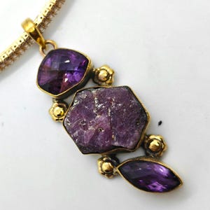 Könnte beinhalten: Eine goldfarbene Anhängerkette mit einem großen, roh geschliffenen, sechseckigen violetten Stein in der Mitte, flankiert von zwei kleineren facettierten violetten Steinen. Die Fassung enthält kleine goldfarbene Blumenakzente und eine Gliederkette.