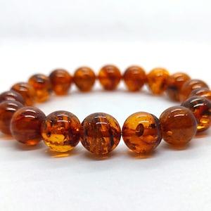 May include: A bracelet featuring polished amber beads. The beads are a warm, translucent brown color with natural inclusions, strung together on an elastic band. The bracelet is displayed on a white surface.