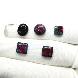 May include: An assortment of polished gemstones in various shapes: oval, round, and square. The stones feature a deep red hue with hints of blue and green. The gemstones are displayed on a white surface.