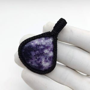 May include: A teardrop-shaped purple and white stone pendant encased in a black macrame cord setting. The stone features a mottled, marbled texture with deep violet and white patches throughout. The black cord forms a loop at the top for hanging.