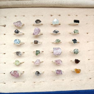 May include: A collection of twenty-four rings featuring raw, uncut gemstones in various colors including pink, blue, green, brown, and black. Each ring has a thin silver-toned metal band holding a unique, irregular mineral specimen on top.