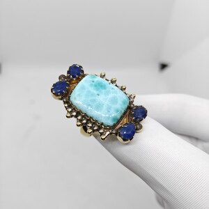 May include: A gold-toned ring featuring a large, rectangular, light blue gemstone, surrounded by smaller, dark blue gemstones. The ring has an ornate, textured setting.