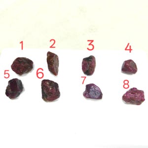 Puede incluir: Se muestran ocho piedras preciosas de rubí en bruto, cada una con un tono rojo intenso. Las piedras varían en tamaño y forma, con texturas naturales visibles y algunas con inclusiones de color verde oscuro. Las piedras preciosas están dispuestas sobre una superficie blanca.