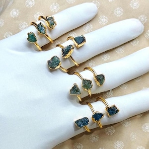 Peut inclure: Une collection de bagues dorées ornées de pierres précieuses brutes et non taillées de couleur sarcelle. Chaque bague possède un anneau métallique fin et réglable avec une monture en pierre brute. Les pierres varient en forme et en taille.