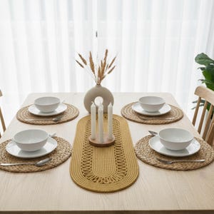 Könnte beinhalten: Ein Esstisch für fünf Personen mit gewebten Tischsets, weißen Schalen und Tellern sowie Besteck. Ein gelber Tischläufer mit dekorativem Muster verläuft in der Mitte. Eine Vase mit getrocknetem Weizen und Kerzen vervollständigen die Szene.