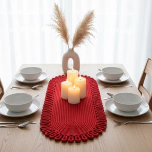 May include: A dining table set for four with a red oval table runner, white dishes, and lit candles. A vase with dried flowers sits in the center. The table is made of light-colored wood.
