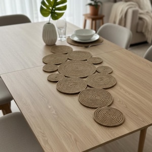 Puede incluir: Una mesa de comedor de madera clara con un juego de manteles individuales y posavasos de yute tejido. Un jarrón blanco con una hoja verde grande y un cuenco blanco sobre un mantel individual también están sobre la mesa.