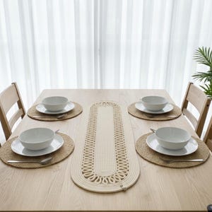May include: A wooden dining table set for four with white bowls, plates, and silverware. The table is adorned with woven placemats and a cream-colored table runner. The room has a bright, airy feel with sheer curtains in the background.