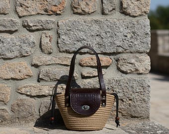Può includere: Una borsa a mano in paglia intrecciata con patta e manici in pelle marrone scuro. La borsa ha una chiusura con coulisse e una chiusura color argento. La borsa è appoggiata su una superficie di pietra davanti a un muro di pietra.
