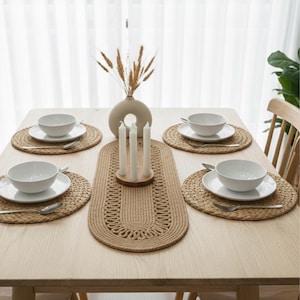 May include: A dining table set for five with woven placemats, white bowls, and silverware. A woven table runner and a vase with dried wheat stalks are centered. White candles in a wooden holder add to the decor.