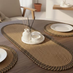 Könnte beinhalten: Ein runder Holztisch mit einem gewebten braun-schwarzen ovalen Tischläufer und passenden Tischsets. Eine weiße Keramikvase, eine Kerze und dekorative Steine ​​liegen auf einem weißen Tablett. Auf einem der Tischsets befindet sich ein weißer Teller.