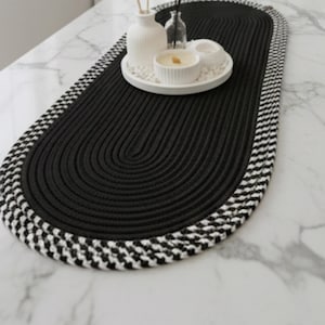 Könnte beinhalten: Ovale schwarze Tischdecke mit geflochtenem Seildesign. Der Rand hat ein schwarz-weißes Karomuster. Ein weißes Tablett mit Kerzen und Vasen steht auf der Tischdecke und schafft eine dekorative Tischdekoration.