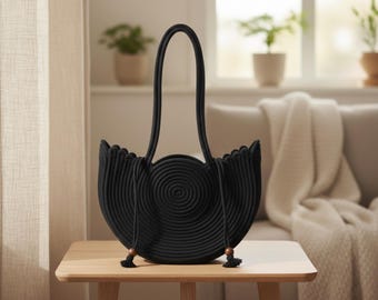 以下が含まれることがあります： 丸いロープで作られた黒いトートバッグが木製のテーブルの上に置かれています。バッグの中央には円形のデザインがあり、縁はスカラップになっています。長いハンドルが上方に伸び、タッセルが下からぶら下がっています。
