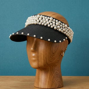 May include: A black visor hat with a woven texture, adorned with faux pearls along the brim and crown. The hat is displayed on a wooden mannequin head against a teal background, showcasing a stylish accessory.