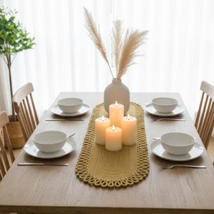 Könnte beinhalten: Ein Holztisch, gedeckt für ein Essen. Ein gelber Tischläufer mit dekorativem Rand liegt in der Mitte. Weiße Schalen, Teller und Besteck sind für vier Personen gedeckt. Angezündete Kerzen und eine Vase mit Ziergras vervollständigen das Arrangement.