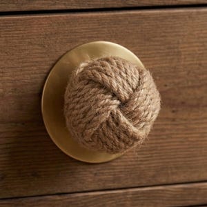 Może przedstawiać: Gałka do szafki z okrągłą, mosiężną podstawą i uchwytem z węzła z liny. Lina ma naturalny, brązowy kolor, a gałka jest zamontowana na drewnianej powierzchni z widocznym usłojeniem.