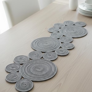 Peut inclure: Un chemin de table gris en corde circulaire avec un motif en spirale. Le chemin de table est composé de plusieurs cercles reliés de tailles variables, disposés en un motif fluide. Le chemin de table est posé sur une table en bois de couleur claire.