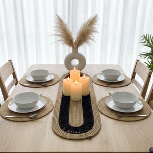 Könnte beinhalten: Ein Esstisch für sechs Personen mit neutralem Farbschema. Der Tisch ist mit weißen Schalen, Tellern und Löffeln gedeckt. Ein gewebter Tischläufer mit schwarzem Design trägt vier brennende Kerzen. Eine Vase mit Trockenblumen steht in der Mitte.