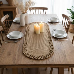 May include: A wooden dining table set for a meal, featuring a woven beige table runner with a decorative edge. White bowls, plates, and silverware are set for each place setting. Three lit candles and a white vase with dried flowers complete the decor.