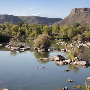 May include: A scenic landscape featuring a calm river with rocky islands and lush green trees. Mountains rise in the background under a clear blue sky, creating a serene natural environment.