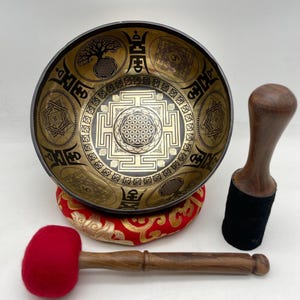 Può includere: Una ciotola tibetana dorata con intricati disegni incisi, tra cui un albero della vita e motivi geometrici. Poggia su un cuscino rosso e oro, con un martello di legno e un battente di feltro rosso.