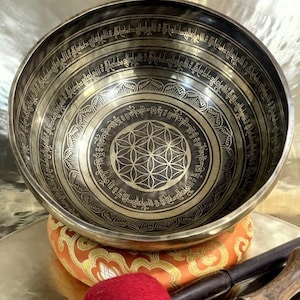 May include: A bronze singing bowl with intricate etched patterns and a central flower of life design. The bowl rests on a patterned cushion, with a red mallet and wooden striker visible.