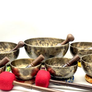 May include: A collection of seven brass singing bowls with a dark, brushed finish and a circular etched symbol on the exterior. Each bowl includes a wooden mallet, and two large mallets with red felt heads are positioned in the foreground.