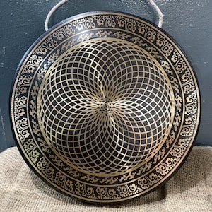 May include: A large, round gong with a black background and gold geometric design. The gong has an intricate pattern in the center and a decorative border. A white rope is attached for hanging.
