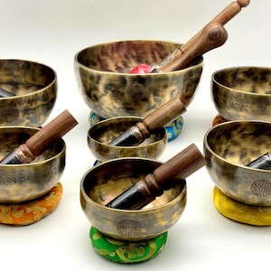 Puede incluir: Conjunto de cuencos tibetanos de bronce con mazos de madera. Los cuencos, de diferentes tamaños, descansan sobre cojines coloridos. Cada cuenco tiene un patrón y diseño únicos, algunos con símbolos y texto grabados.