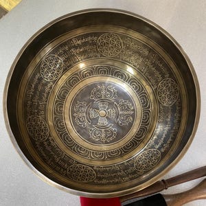 Può includere: Una ciotola tibetana color bronzo con intricati motivi incisi. La ciotola presenta un disegno centrale circondato da motivi floreali e geometrici e da una scritta. Il bordo presenta una serie di motivi ripetuti. La ciotola è progettata per la guarigione del suono e la meditazione.