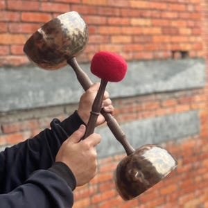 Può includere: Due ciotole tibetane con manici in legno e un mazzuolo rosso sono tenute davanti a un muro di mattoni. Le ciotole hanno una finitura metallica e il mazzuolo ha una testa rossa arrotondata. I manici sono marrone scuro.
