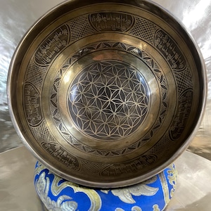 Può includere: Una ciotola tibetana color bronzo con intricati motivi incisi e un disegno geometrico al centro. La ciotola poggia su un cuscino blu e oro, creando un contrasto sorprendente. Il bordo della ciotola presenta una scritta incisa.