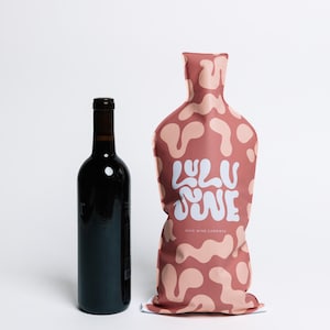 May include: A dark wine bottle stands next to a wine carrier. The carrier is a dusty rose color with a pattern of light pink organic shapes. The text "Lulu Wine" is printed on the carrier in white, with "Chic Wine Carriers" below.