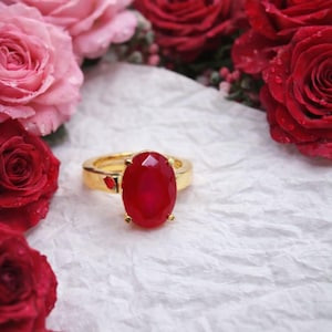 Op de afbeelding: Een gouden ring met een grote, ovale, rode edelsteen. De ring staat tegen een achtergrond van rode en roze rozen op een wit, getextureerd oppervlak. De band van de ring heeft een klein rood accent.