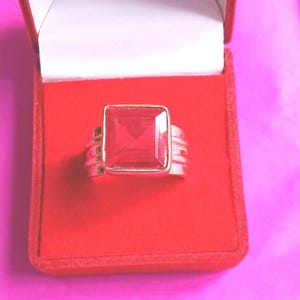 Op de afbeelding: Een goudkleurige ring met een grote, rechthoekige, rode edelsteen. De ring wordt gepresenteerd in een rode fluwelen doos met een witte binnenkant, tegen een roze achtergrond. De edelsteen is ongeveer 2 cm lang.